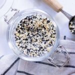 A glass jar filled with everything bagel seasoning sits on a striped cloth, with a wooden-handled measuring spoon nearby.