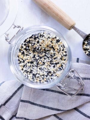 A glass jar filled with everything bagel seasoning sits on a striped cloth, with a wooden-handled measuring spoon nearby.