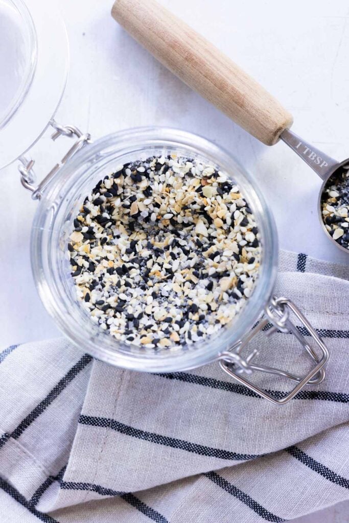 A glass jar filled with everything bagel seasoning sits on a striped cloth, with a wooden-handled measuring spoon nearby.