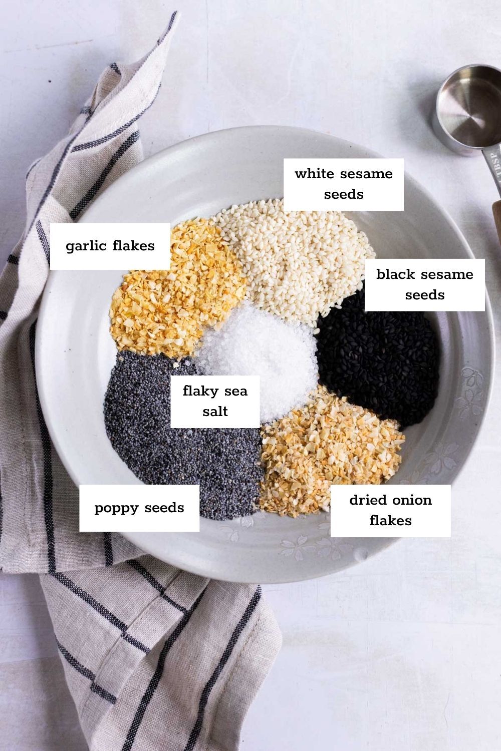 A white bowl contains separate piles of garlic flakes, white sesame seeds, black sesame seeds, flaky sea salt, poppy seeds, and dried onion flakes, each labeled.
