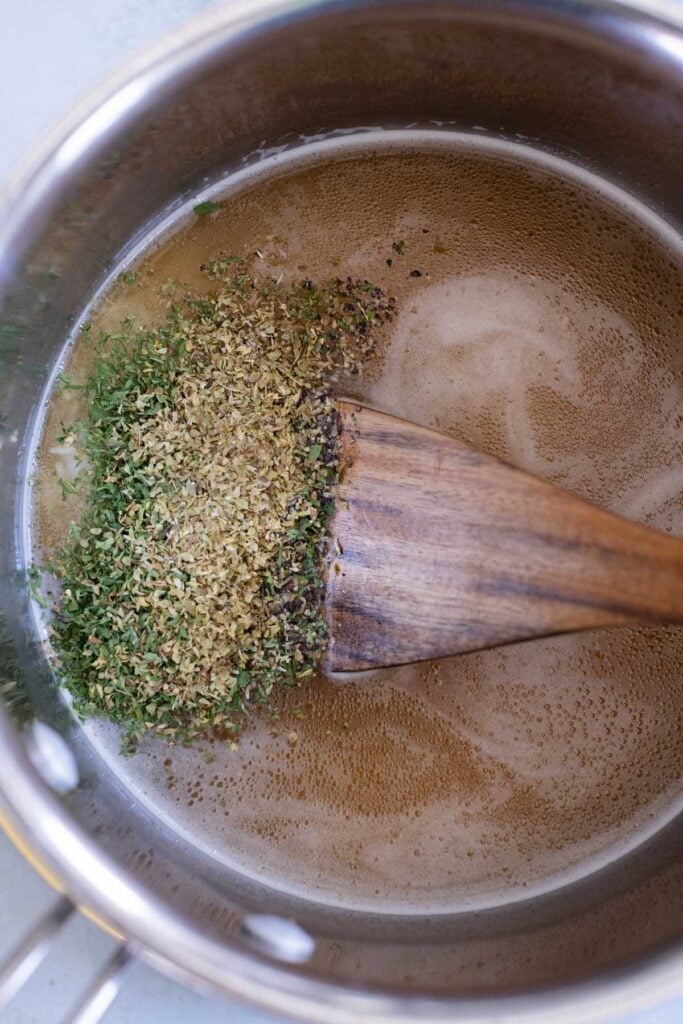 A metal saucepan containing brown liquid and mixed dried herbs being stirred with a wooden spatula.