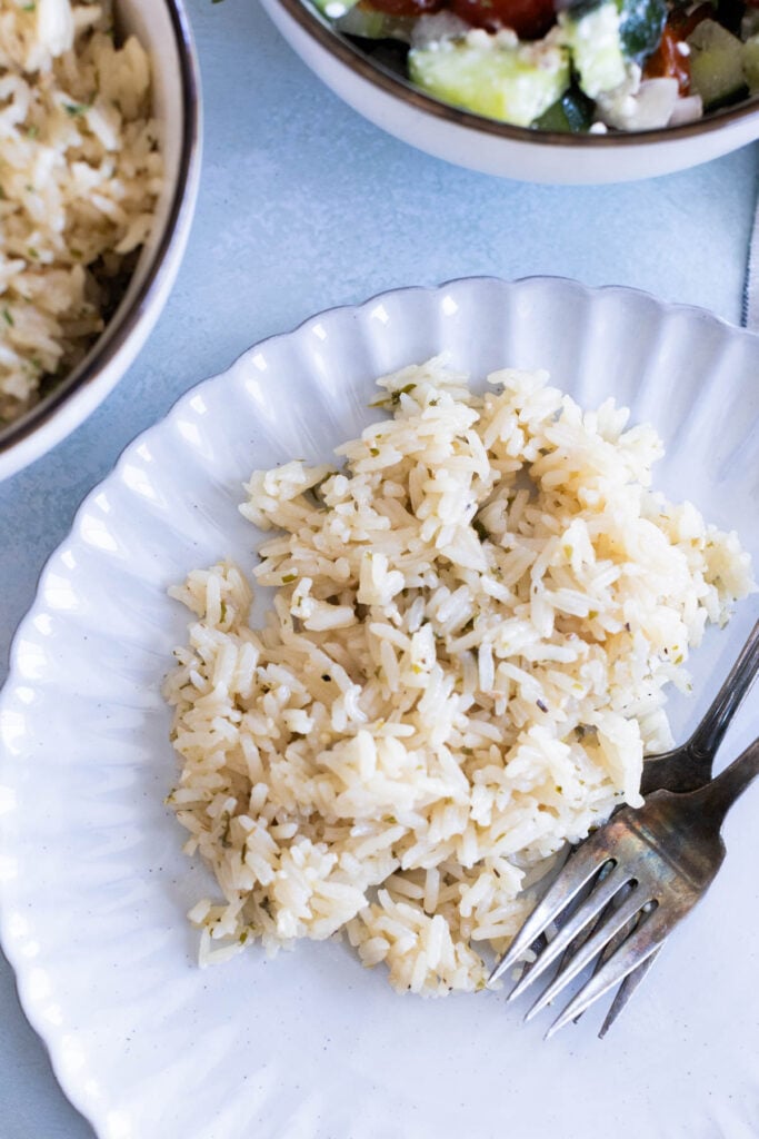 A serving of greek white rice on a white plate with a fork and spoon, with bowls of rice and salad nearby.