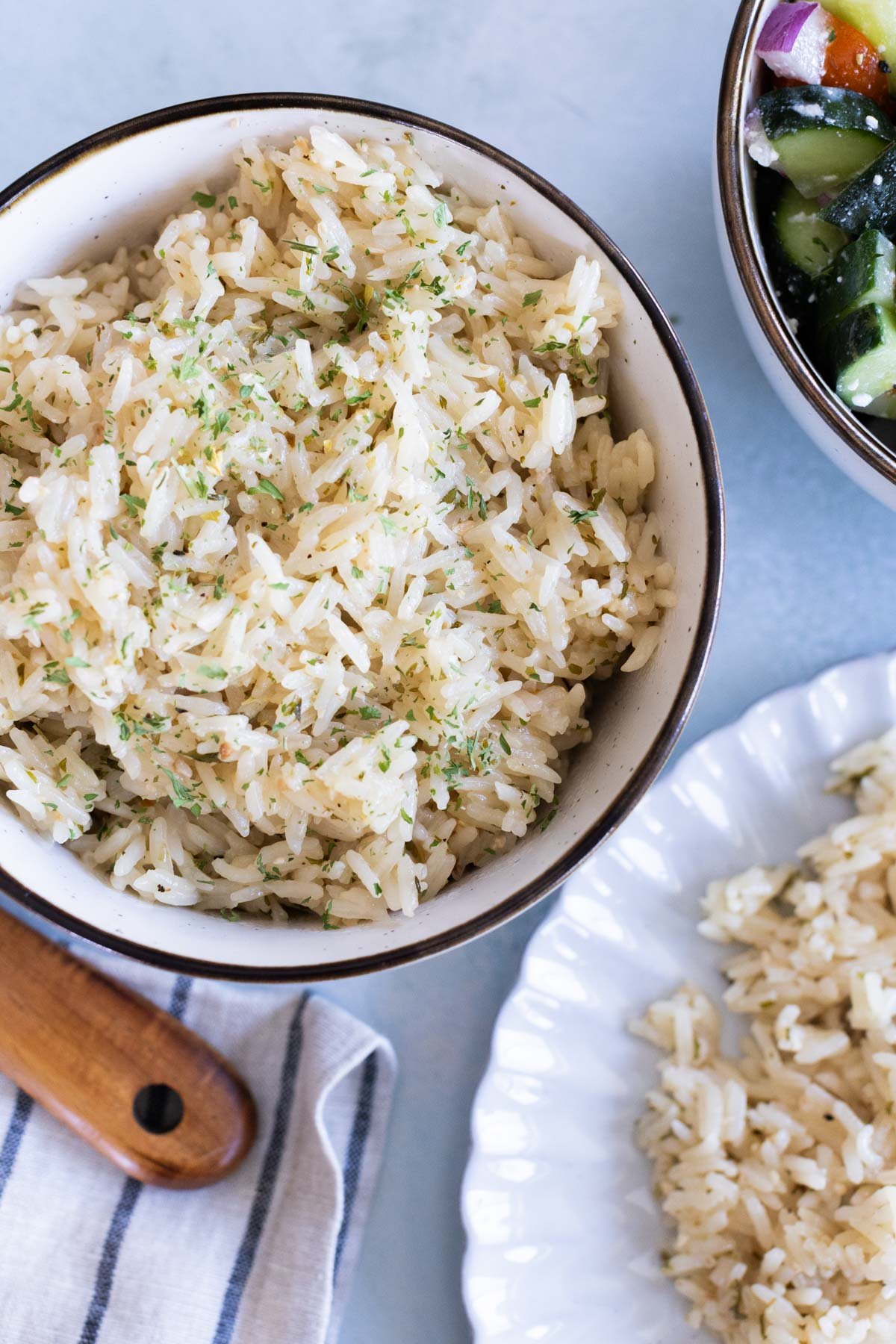 A bowl of cooked, greek rice garnished with herbs sits next to a plate with more rice and part of a salad in a separate bowl.