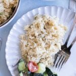 A white plate with a serving of greek rice and a small portion of cucumber and tomato salad, with a fork and knife on the side.
