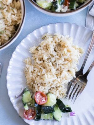 A white plate with a serving of greek rice and a small portion of cucumber and tomato salad, with a fork and knife on the side.