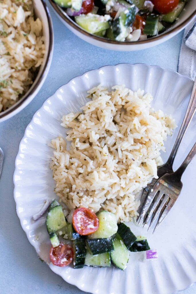 A white plate with a serving of greek rice and a small portion of cucumber and tomato salad, with a fork and knife on the side.