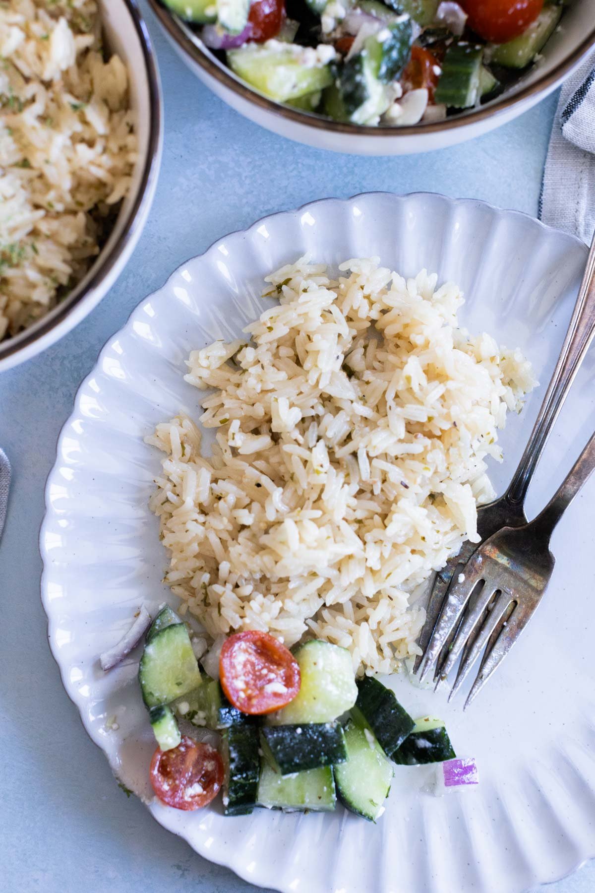 A white plate with a serving of greek rice and a small portion of cucumber and tomato salad, with a fork and knife on the side.
