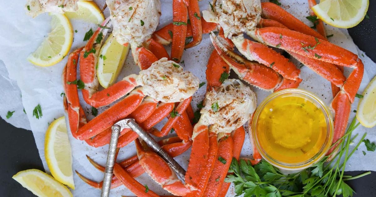 Cooked crab legs garnished with parsley, served with lemon wedges and a small bowl of melted butter on parchment paper, alongside seafood crackers.