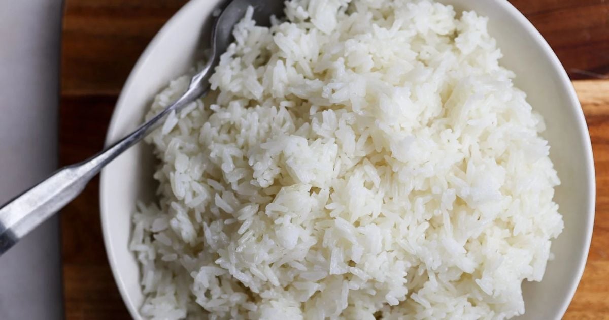 A bowl of cooked white rice with a metal spoon resting on the edge, placed on a wooden surface.