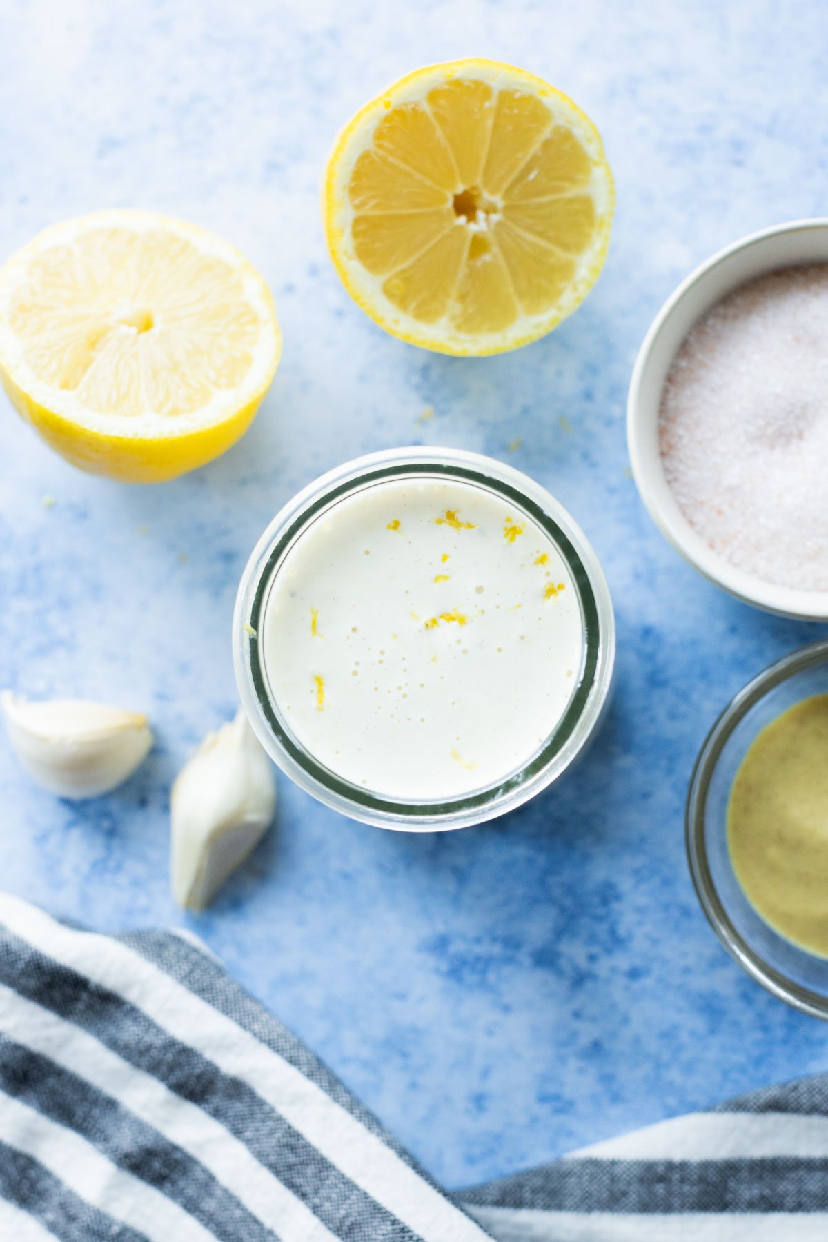 A jar of creamy lemon aioli surrounded by lemon halves, a bowl of pink salt, two garlic cloves, a bowl of mustard, and a striped cloth on a blue surface.