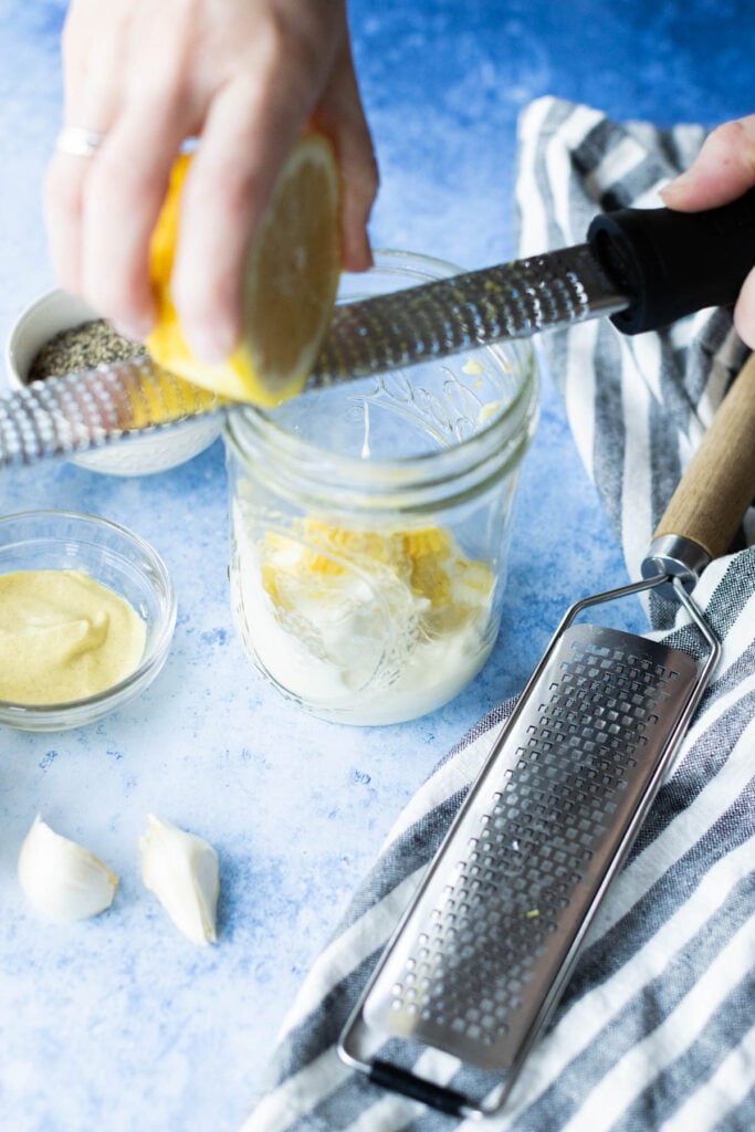 A hand grates a lemon over a glass jar containing grated lemon peel, with mustard, garlic cloves, and a grater nearby on a blue surface with a striped cloth.