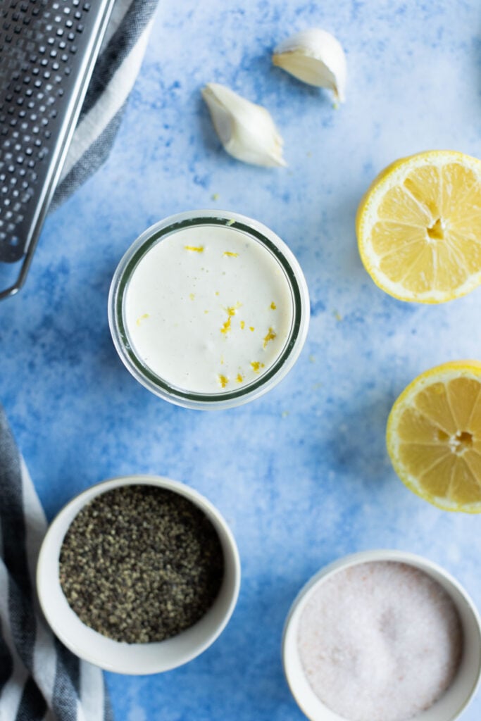 A jar of creamy lemon aioli sits on a blue surface surrounded by halved lemons, garlic cloves, ground black pepper, salt bowls, and a metal grater.