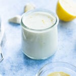 A small glass jar filled with creamy white lemon aioli sits on a blue surface, with half a lemon, garlic cloves, and a bowl of mustard nearby.