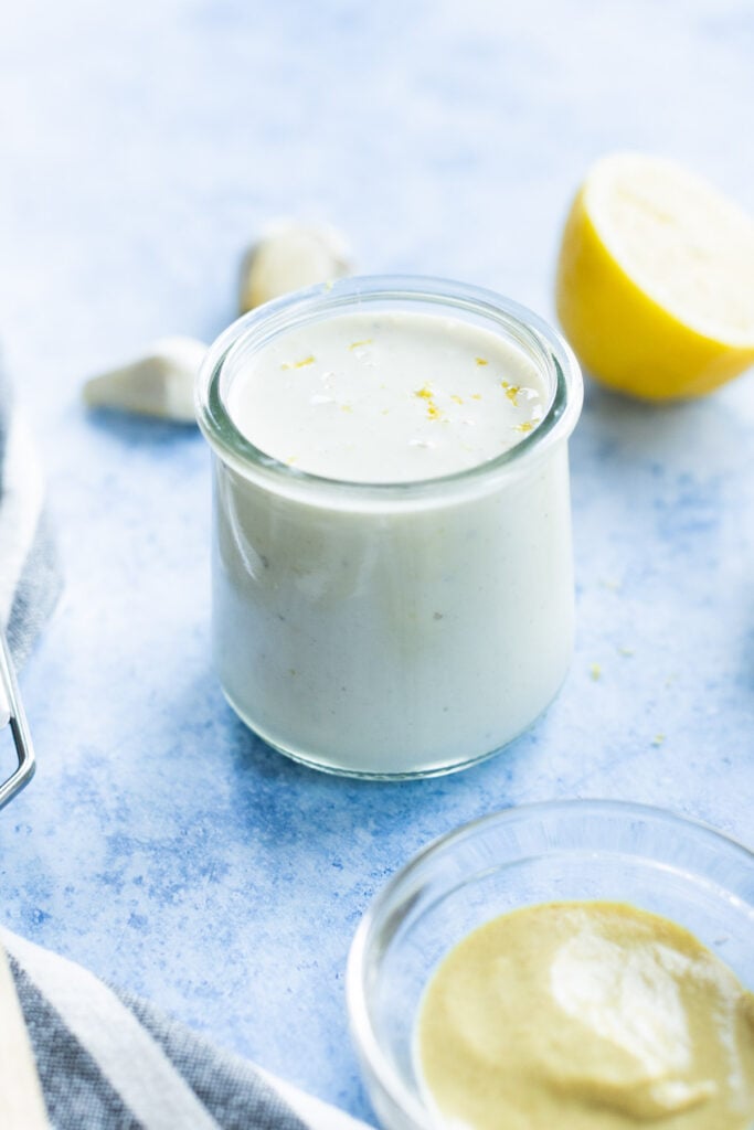 A small glass jar filled with creamy white lemon aioli sits on a blue surface, with half a lemon, garlic cloves, and a bowl of mustard nearby.