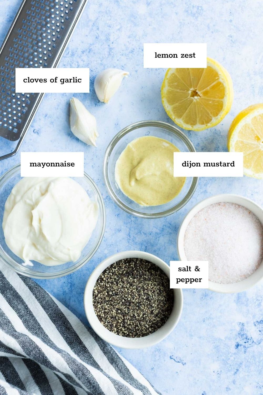 A flat lay of ingredients for lemon aioli, including garlic cloves, lemon zest, mayonnaise, Dijon mustard, salt, and pepper, each labeled in small bowls on a blue surface with a striped cloth.