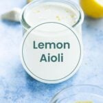 A glass jar of lemon aioli sits on a light blue surface, ready to be paired with air fryer potato wedges. A lemon half and garlic cloves are in the background. Text on the jar reads "Lemon Aioli.