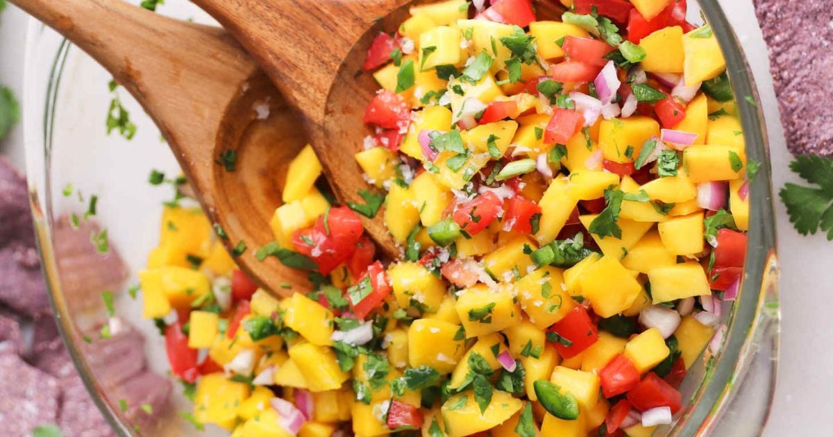 A glass bowl of mango salsa with diced mango, tomato, red onion, cilantro, and lime, mixed with two wooden spoons.