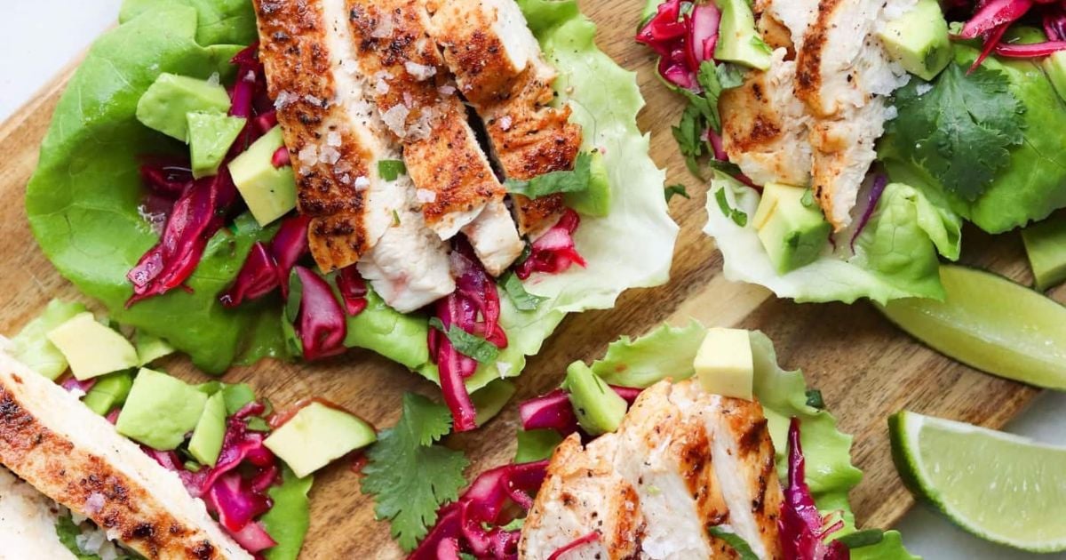 Grilled chicken strips on lettuce leaves, topped with avocado cubes, red cabbage, cilantro, and served with lime wedges on a wooden board.
