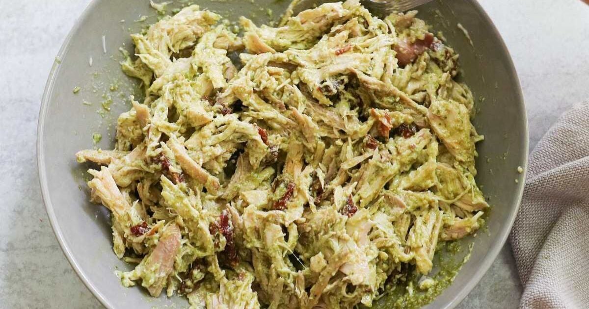 Shredded chicken mixed with green pesto sauce and pieces of sun-dried tomatoes in a gray bowl.
