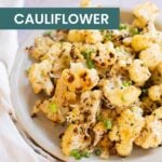 A plate of roasted cauliflower garnished with chopped green onions sits on a white surface with a light-colored cloth nearby. The text overlay reads "roasted CAULIFLOWER," highlighting this delicious vegetable dish.