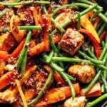 A skillet with cooked chicken pieces, green beans, red bell peppers, and carrot strips in a brown sauce, topped with sesame seeds.