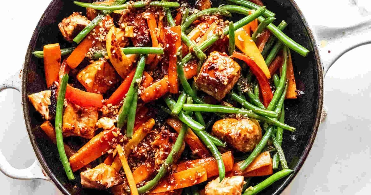 A skillet with cooked chicken pieces, green beans, red bell peppers, and carrot strips in a brown sauce, topped with sesame seeds.