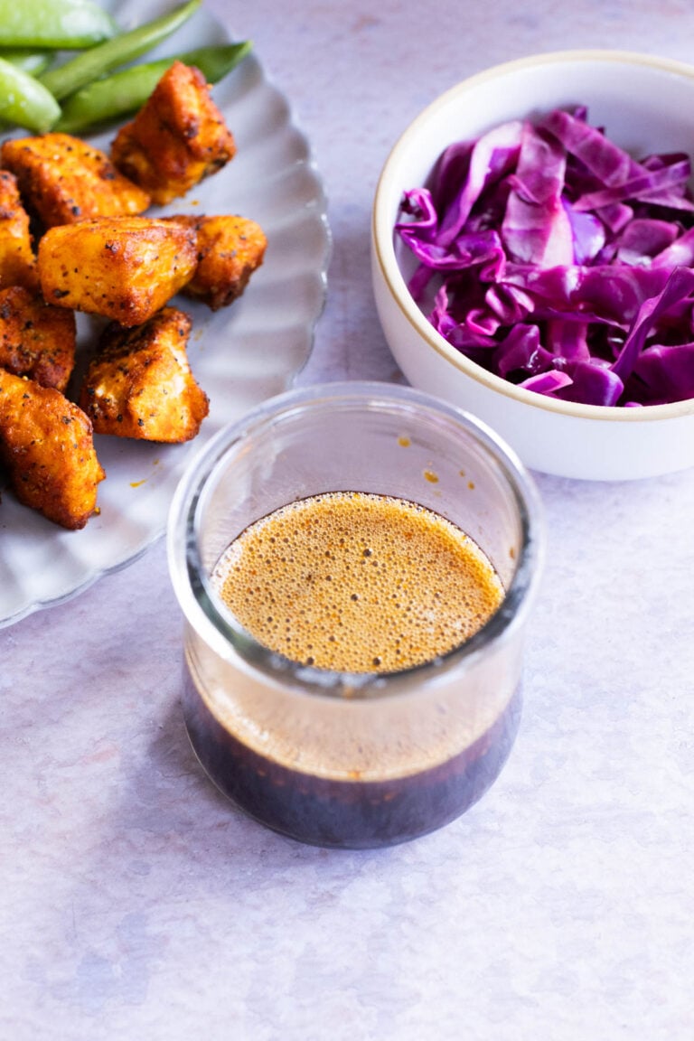 A stir fry sauce in a small, glass jar. A small white bowl contains red cabbage. Salmon bites sit on a plate with fresh green beans.