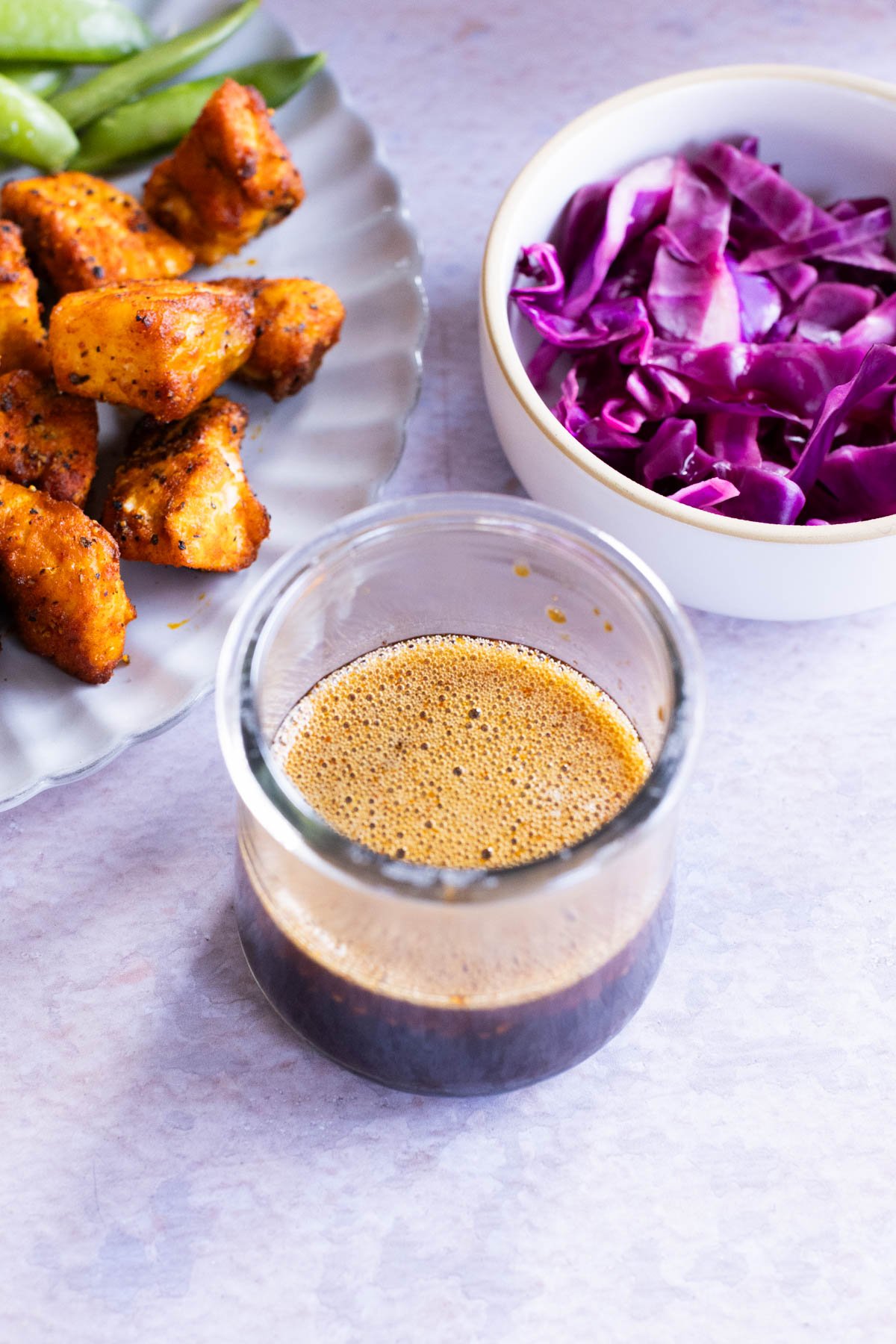 A stir fry sauce in a small, glass jar. A small white bowl contains red cabbage. Salmon bites sit on a plate with fresh green beans.