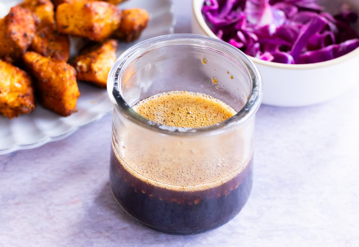 A glass jar filled with a stir fry sauce sits on on a light surface, with a bowl of shredded red cabbage and a plate of seasoned chicken pieces in the background.