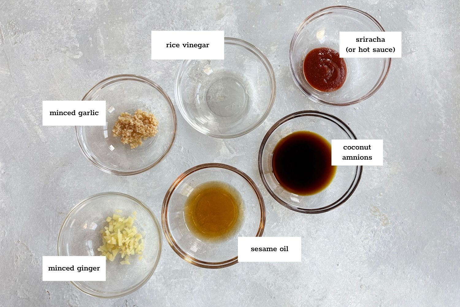 Six small glass bowls containing minced garlic, minced ginger, rice vinegar, sesame oil, coconut aminos, and sriracha, each labeled on a light gray surface.