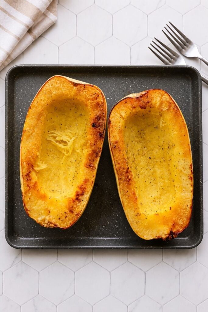 Two roasted spaghetti squash halves on a baking tray, seasoned with salt and pepper.