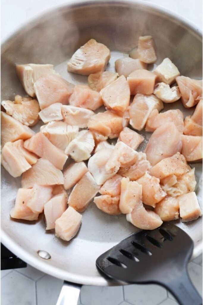 Cubes of boneless chicken breast are being cooked in a stainless steel skillet, with a black spatula.