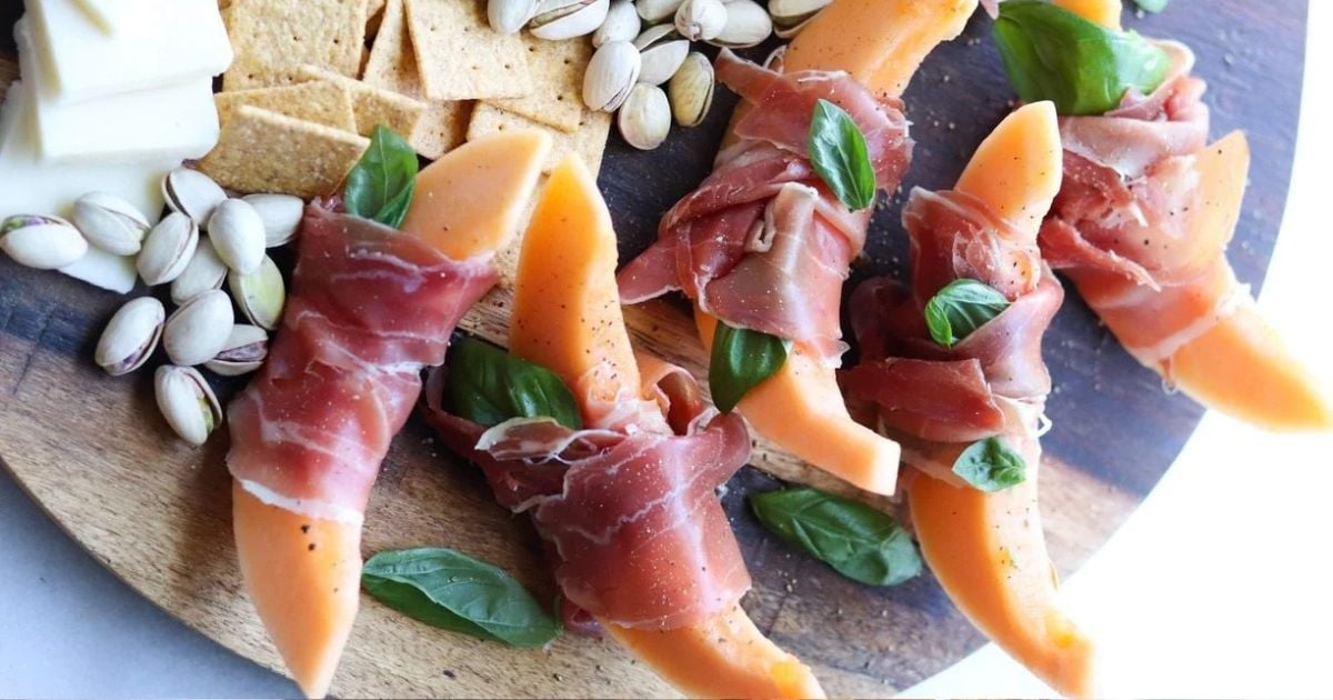 Sliced cantaloupe wrapped in prosciutto with basil leaves, served on a wooden board with crackers, pistachios, and cheese.