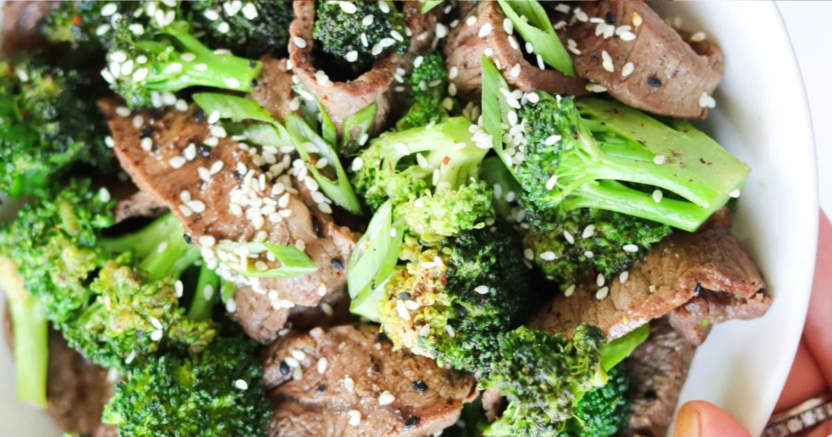 A close-up of a dish featuring sliced beef, broccoli florets, and white sesame seeds in a white bowl.