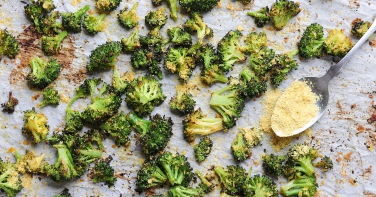 Roasted broccoli florets on a parchment-lined baking sheet with a spoon holding nutritional yeast sprinkled over some pieces.