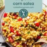 A bowl of chipotle corn salsa with corn, tomatoes, red onion, and cilantro sits beside herbs and lime—perfect as a fresh topping for oven baked salmon in foil.