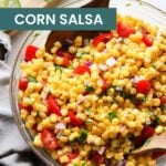 A clear glass bowl filled with corn salsa—corn, diced tomatoes, red onion, and cilantro—sits on a light surface with a gray napkin nearby, perfect as a fresh topping for oven baked salmon in foil.