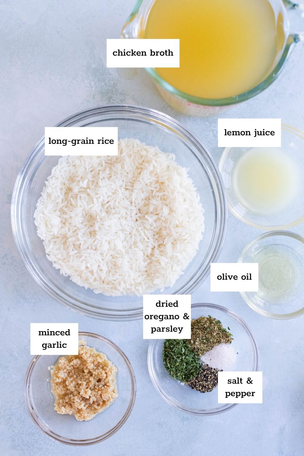 Top-down view of labeled ingredients for a greek rice recipe, including chicken broth, long-grain rice, lemon juice, olive oil, minced garlic, dried oregano and parsley, salt, and pepper.