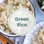 A bowl of cooked Greek rice garnished with herbs, a side plate of greek rice, and a bowl of cucumber salad are arranged on a table with a striped napkin and wooden spoon.