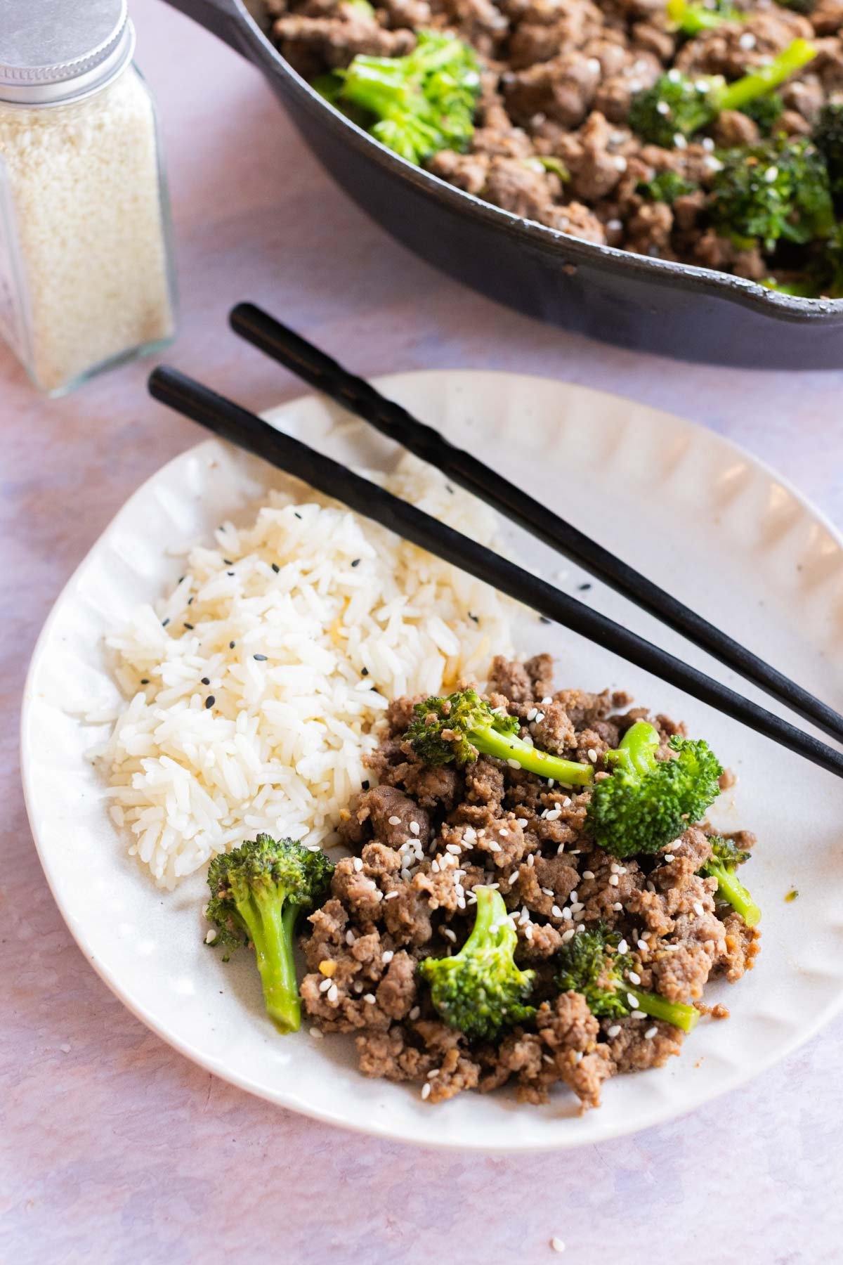 Ground beef with broccoli and rice on a plate with black chopsticks, with a skillet of the same dish and a shaker of sesame seeds in the background.