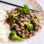 A plate with cooked ground beef, broccoli florets, steamed white rice, and a sprinkle of sesame seeds, with black chopsticks on the side.