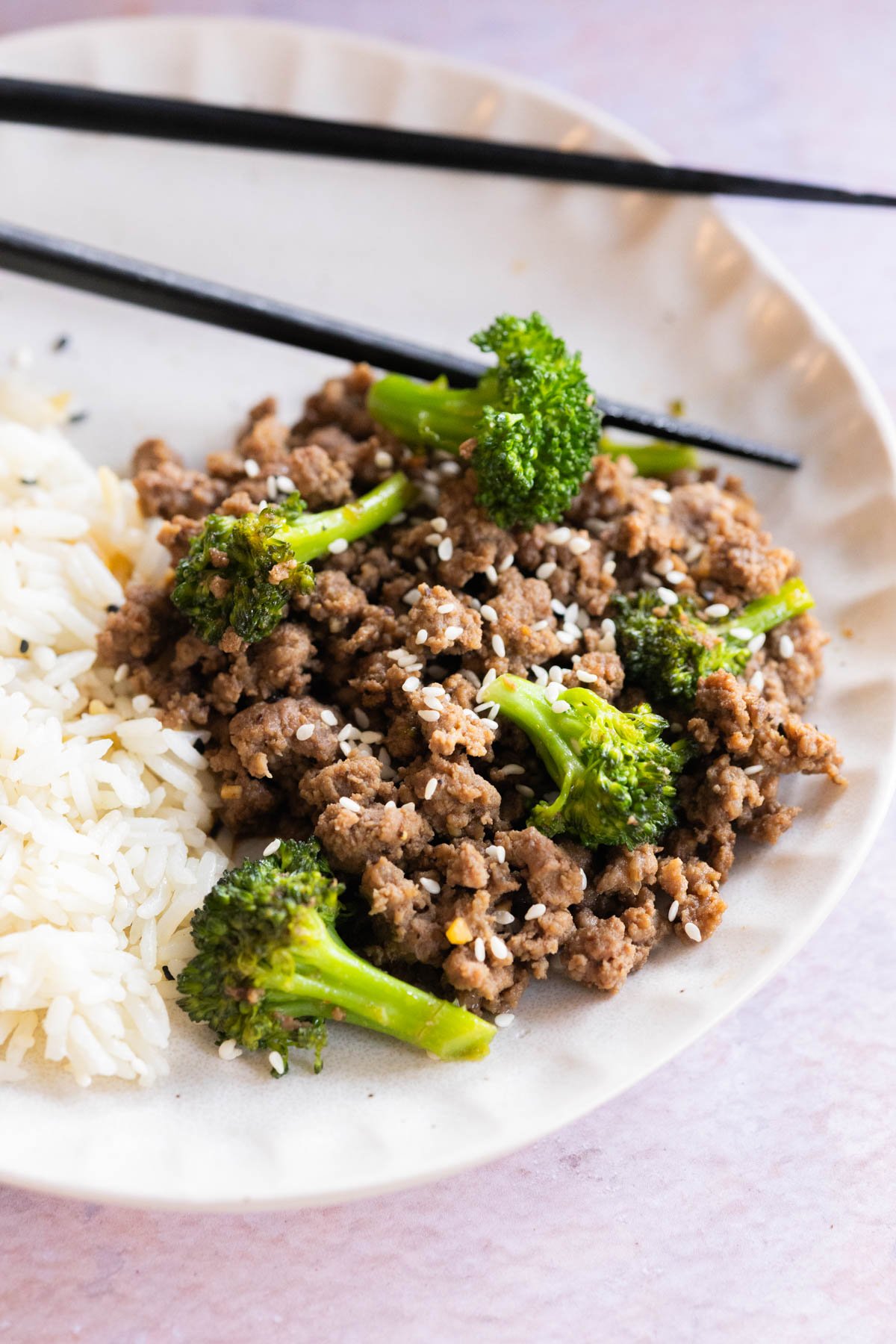 A plate with cooked ground beef, broccoli florets, steamed white rice, and a sprinkle of sesame seeds, with black chopsticks on the side.