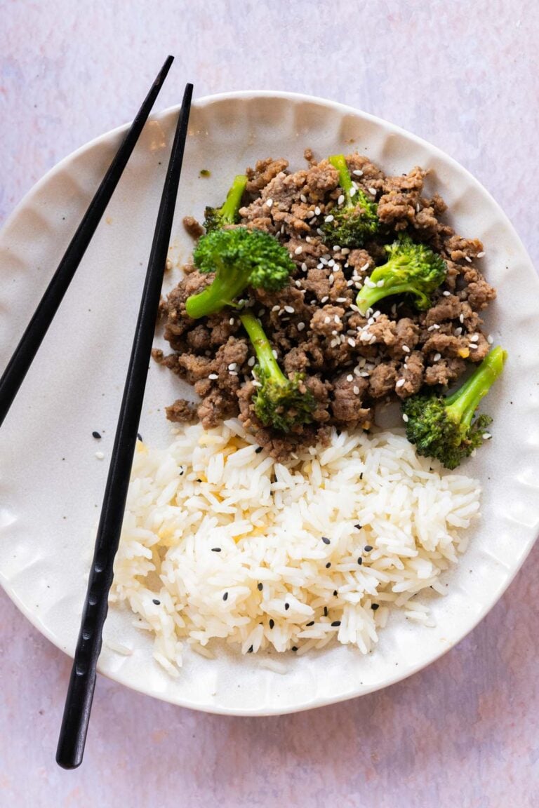 A plate with white rice, ground beef and broccoli topped with sesame seeds, and a pair of black chopsticks on the side.