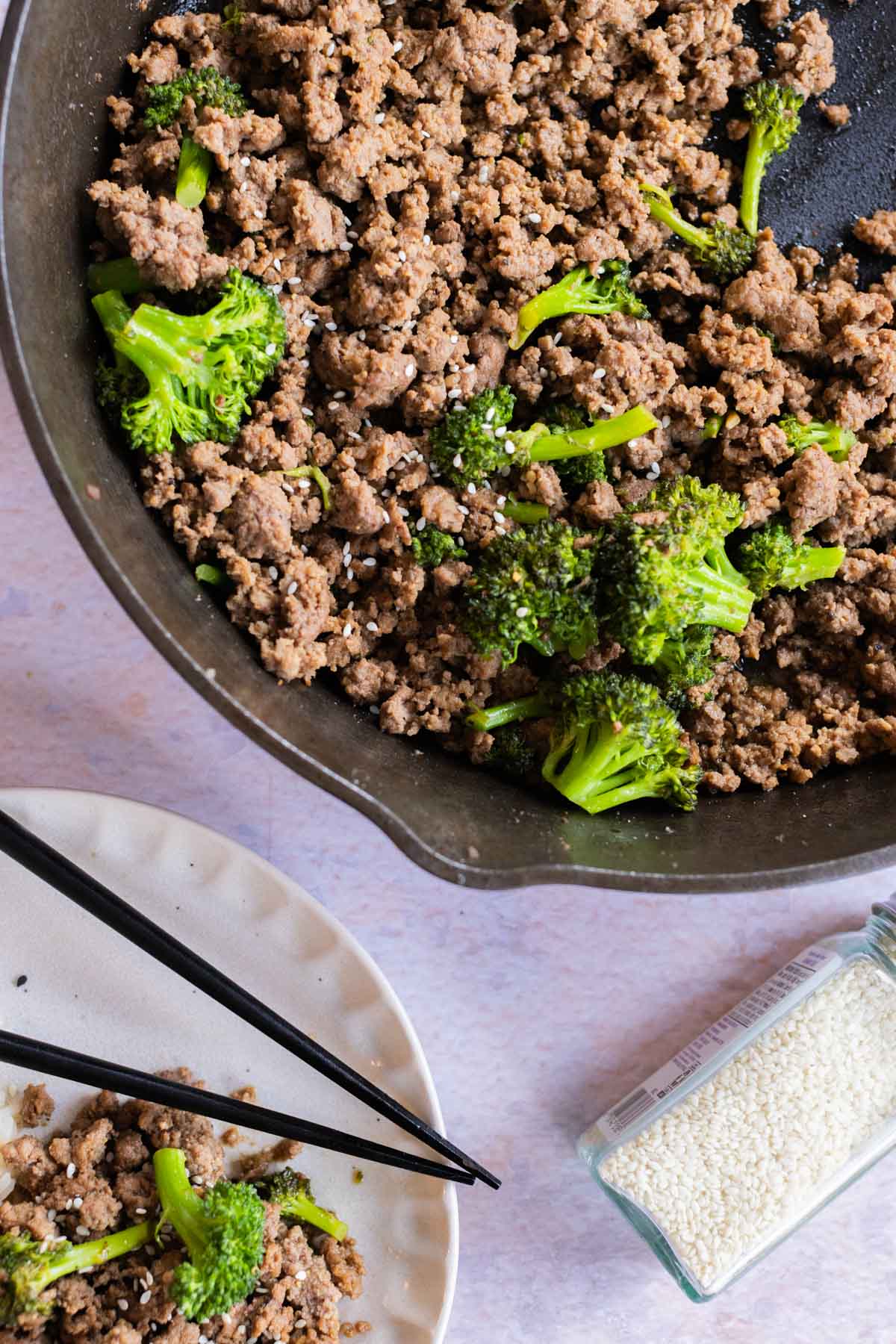 Ground beef and broccoli cooked in a skillet, with sesame seeds sprinkled on top. A plate with chopsticks and a bottle of sesame seeds are next to the skillet.