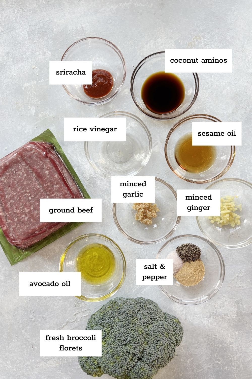 Overhead view of labeled ingredients for a ground beef and broccoli recipe, including garlic, ginger, sriracha, coconut aminos, rice vinegar, sesame oil, avocado oil, salt, and pepper.