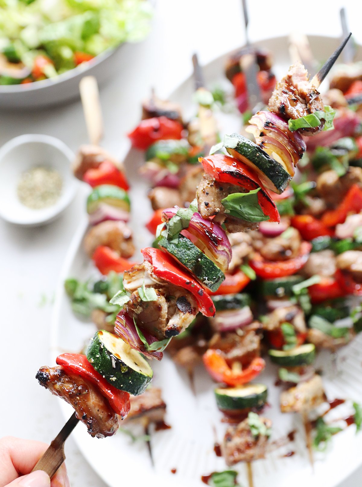 A hand holds a skewer of grilled pork kabobs, featuring juicy meat and vegetables like zucchini, red bell pepper, and onion, over a plate of similar skewers.