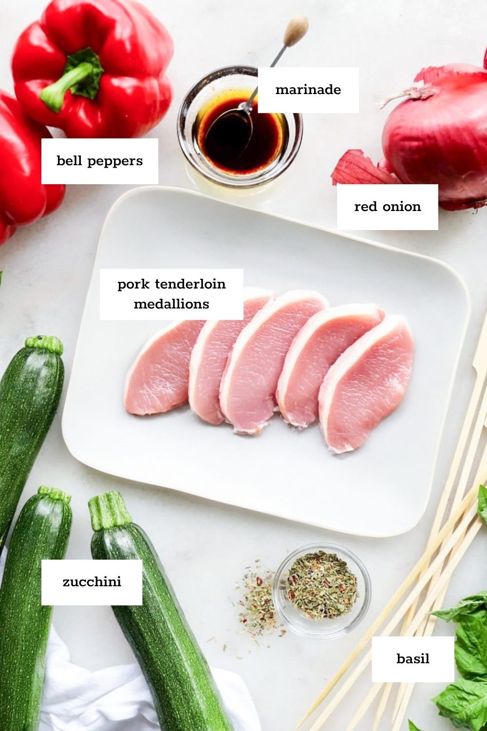 A plate with five pork tenderloin medallions—perfect for making pork kabobs—surrounded by zucchini, bell peppers, red onion, basil, marinade in a bowl, dried herbs, and wooden skewers.
