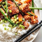 A plate of white rice topped with stir-fried tofu, green beans, carrots, sesame seeds, and chopped green onions, reminiscent of a vibrant salmon stir fry, with black chopsticks on the side.