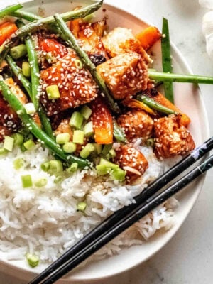 A plate of white rice topped with stir-fried tofu, green beans, carrots, sesame seeds, and chopped green onions, reminiscent of a vibrant salmon stir fry, with black chopsticks on the side.