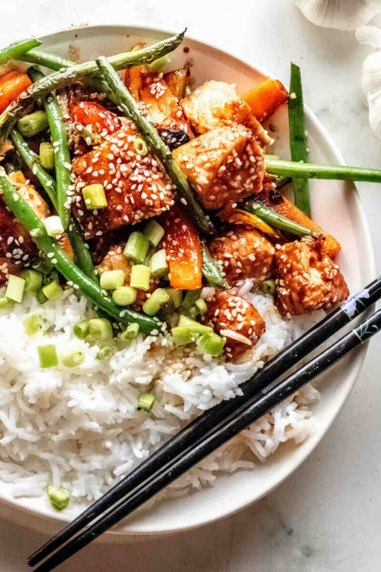 A plate of white rice topped with stir-fried tofu, green beans, carrots, sesame seeds, and chopped green onions, reminiscent of a vibrant salmon stir fry, with black chopsticks on the side.